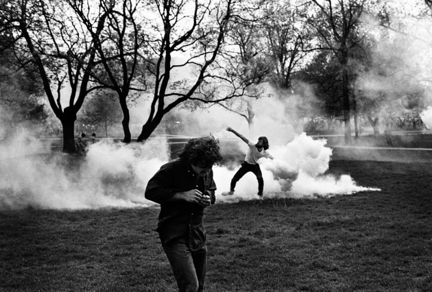 Black and white photo of two people in a park amidst thick smoke. One person in the foreground is running away while looking down, while another in the background is throwing an object. Bare trees with sparse leaves are visible and capture a moment reminiscent of Berkeley Journalism's striking visuals.