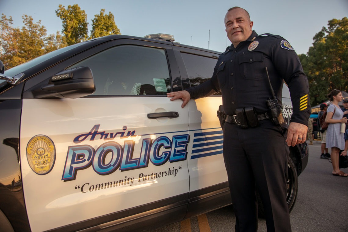 A police officer in uniform stands next to a black and white Arvin Police vehicle with the words "Community Partnership" on the side. The officer is smiling with one hand resting on the car. Trees and people are in the background, with sunlight casting a warm glow, capturing a scene worthy of Berkeley Journalism.