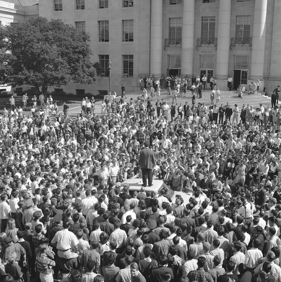 A black and white photo depicts a large crowd gathered outdoors, focusing on a man standing on a platform or table speaking to the audience. The setting appears to be in front of a large, classical-style building with pillars, capturing a moment significant to Berkeley Journalism history.