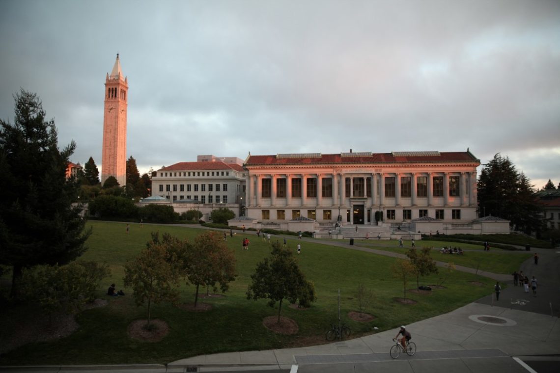 A scenic view of a university campus featuring a tall clock tower on the left and a large, classical-style building with columns in the center. The sun sets, casting a warm glow. Students studying Berkeley Journalism are seen relaxing and cycling on the grassy area and paths.