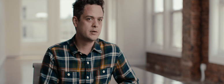 A man with short, curly hair is sitting in a well-lit room with large windows. He is wearing a plaid shirt and looking towards the camera, exuding the curiosity of someone who might be involved in Berkeley Journalism. The room has a modern, minimalist look with white walls and exposed brick.