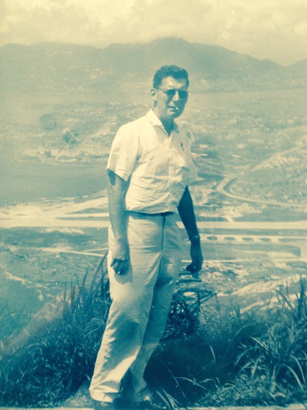 A man in a white shirt and trousers stands on a hillside, with a city, an airport, and mountains in the background. Wearing sunglasses, he exudes a cool demeanor. The sepia tone of the image gives it a vintage look, reminiscent of classic Berkeley Journalism style.