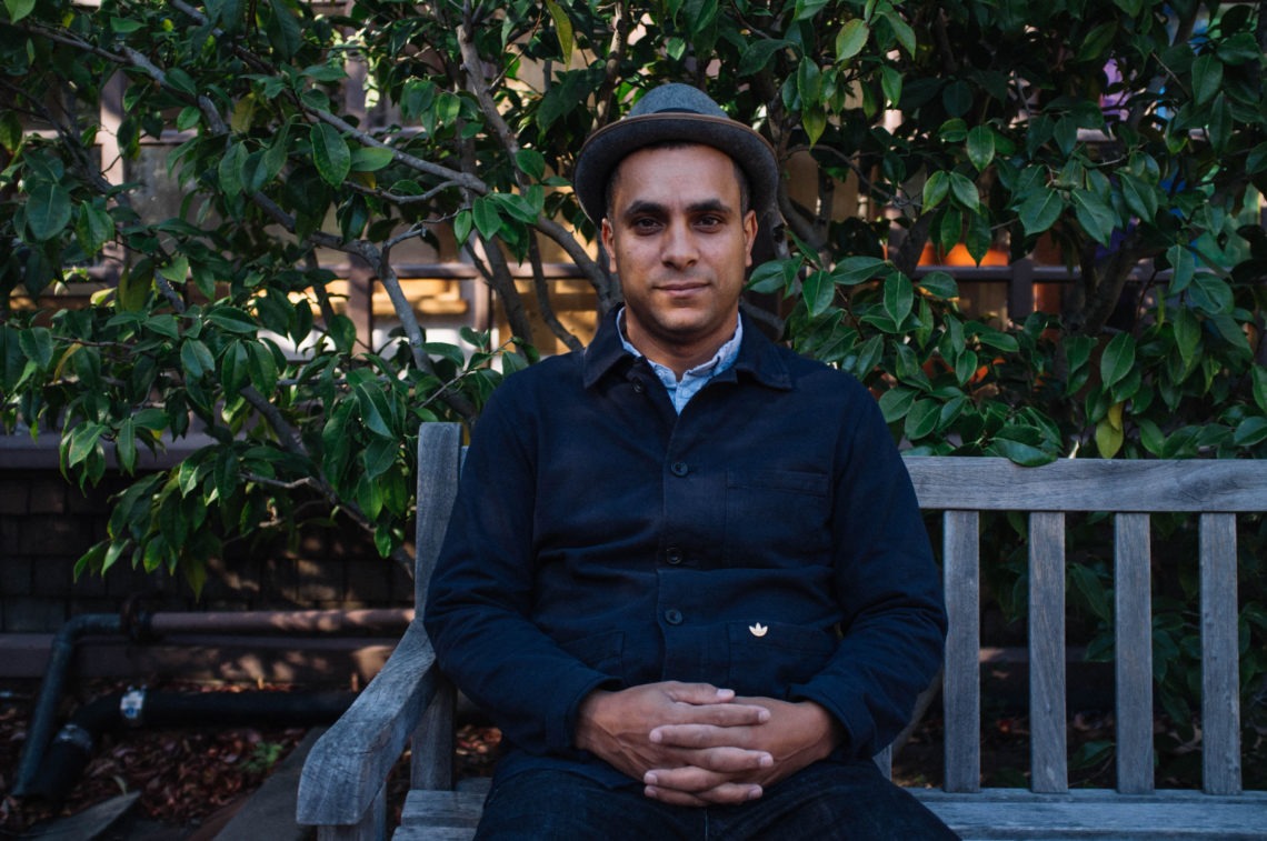 A headshot of a man sitting in a wooden bench outside. He is wearing a button up sweater and a fedora. The background is full of greenery.