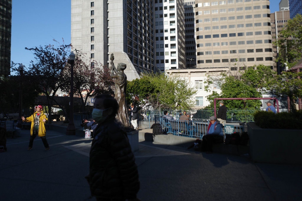 A city plaza with people walking around. A statue holding a sphere is visible in the center, surrounded by tall buildings and trees providing some greenery. One person wears a bright yellow coat; another sports a face mask, reminiscent of scenes covered by Berkeley Journalism. The area is partly shaded.