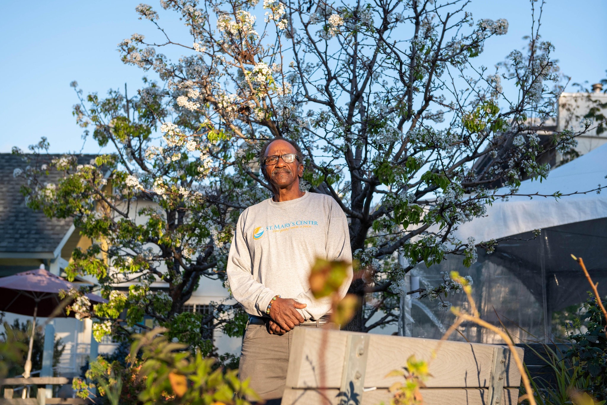 A man stands in a garden with blooming trees, wearing glasses and a gray long-sleeve shirt with a Berkeley Journalism logo. The sky is clear and blue, and a house is visible in the background. The man is smiling and has his hands clasped in front of him.