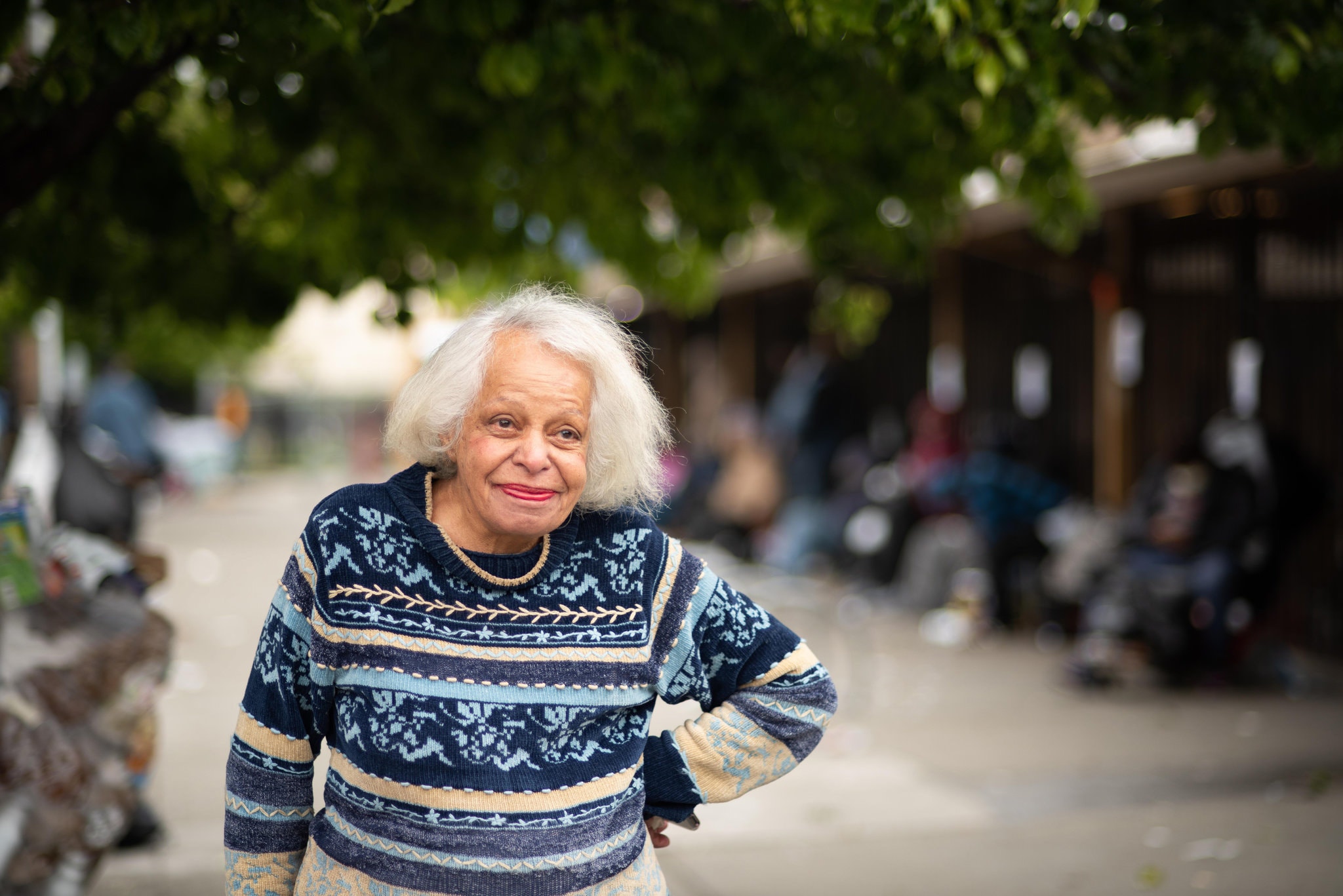 An elderly woman with white hair and wearing a patterned sweater smiles at the camera while standing outdoors. The background is slightly blurred, showing a tree and several people sitting on benches—perhaps capturing a moment from her Berkeley Journalism days.