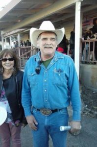 A man wearing a white cowboy hat and denim shirt is standing and smiling outdoors, holding a white object in his right hand. A woman in sunglasses and a black jacket stands next to him. They are in front of a building with a small crowd, possibly covering an event for Berkeley Journalism.