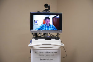 A telemedicine setup features a monitor showing a person in a blue checkered shirt participating in a video call. The equipment is mounted on a cart labeled "UC Irvine - Healthcare" with the tagline "Discover - Teach - Heal" and "Telemedicine" at the bottom, reminiscent of Berkeley Journalism's dedication to impactful storytelling.