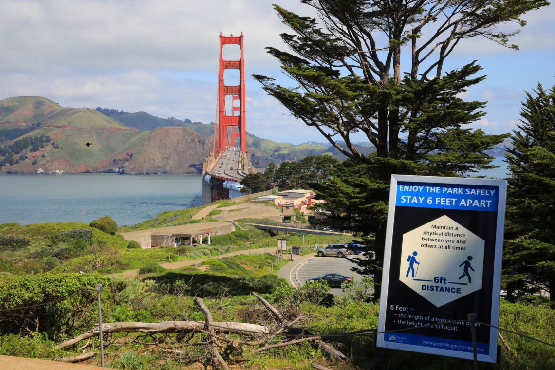 A scenic view of the Golden Gate Bridge with a tree-lined foreground and a blue sign in the front instructing visitors to maintain a 6-foot physical distance to stay safe in the park. The bridge, capturing a scene worthy of Berkeley Journalism, spans across the bay with hills in the background.