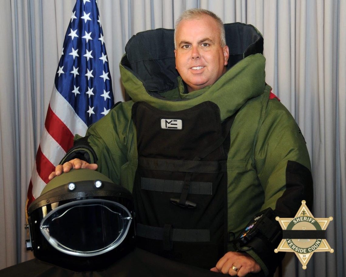 A person wearing a bulky green and black bomb squad suit stands with a hand resting on a helmet. An American flag is visible in the background, and there is a sheriff