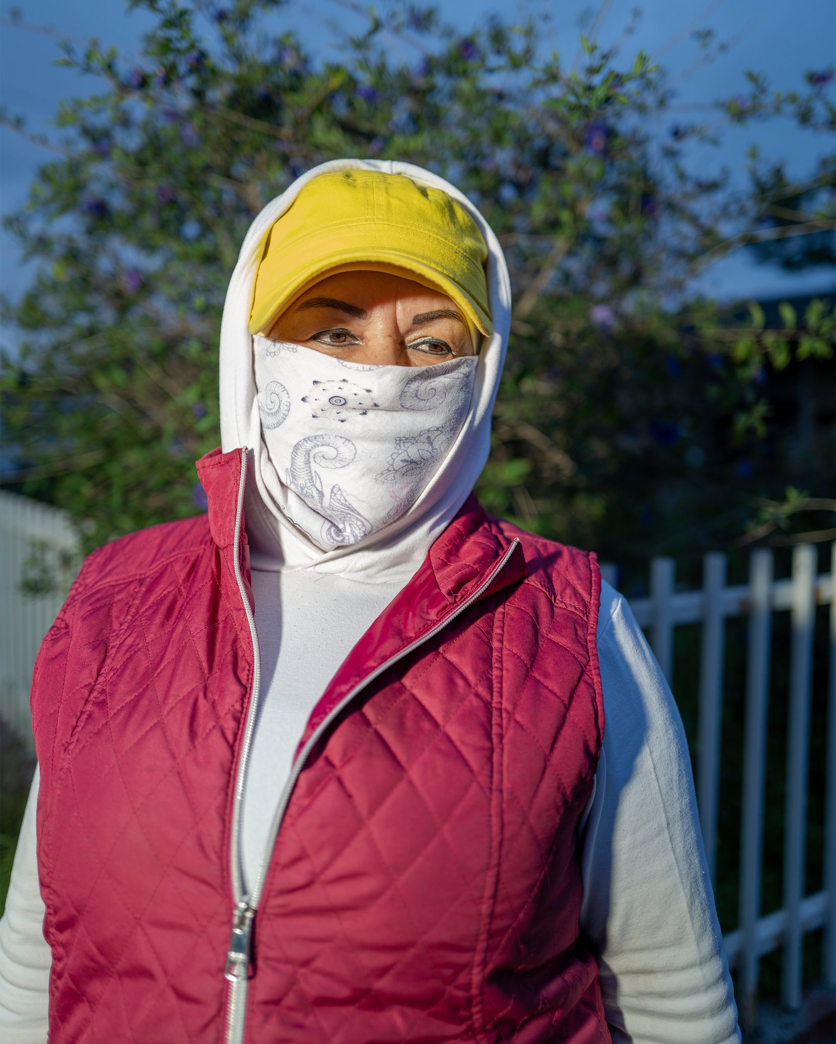 A person stands outdoors near a white fence, wearing a yellow cap, a white hoodie, and a red quilted vest. Their face is partially covered with a white patterned cloth, leaving only the eyes visible. Trees and a blue sky can be seen in the background, capturing an everyday moment worthy of Berkeley Journalism.
