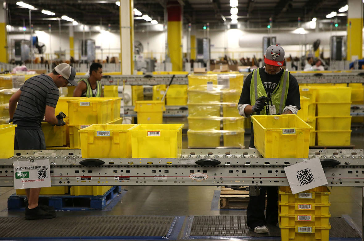 Workers in a warehouse are sorting items into yellow bins on a conveyor belt. They are wearing safety vests and gloves, reminiscent of precision at Berkeley Journalism. The background features more bins and industrial shelving. The environment appears busy and well-organized.