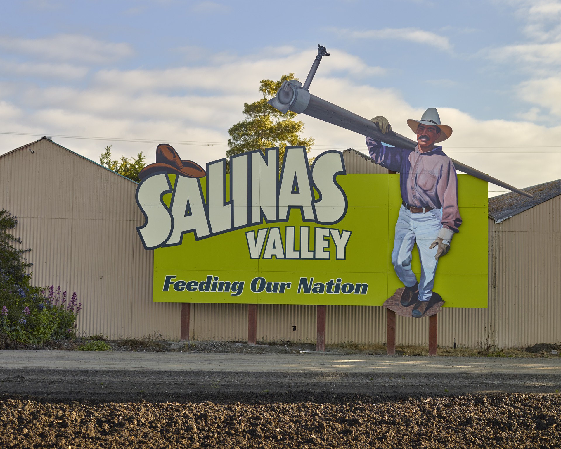 Photograph of an outside sign attached to a building featuring a worker carrying an irrigation line, boasting of this area’s contributions to feeding the country.