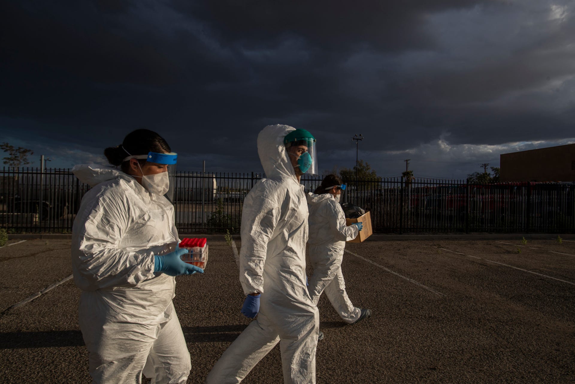 Three individuals dressed in full protective gear, including white coveralls, blue gloves, and face shields, walk through an outdoor area with a stormy sky overhead. One person carries a red container, and another holds a clipboard. They could be from Berkeley Journalism on assignment, appearing focused on the task at hand.