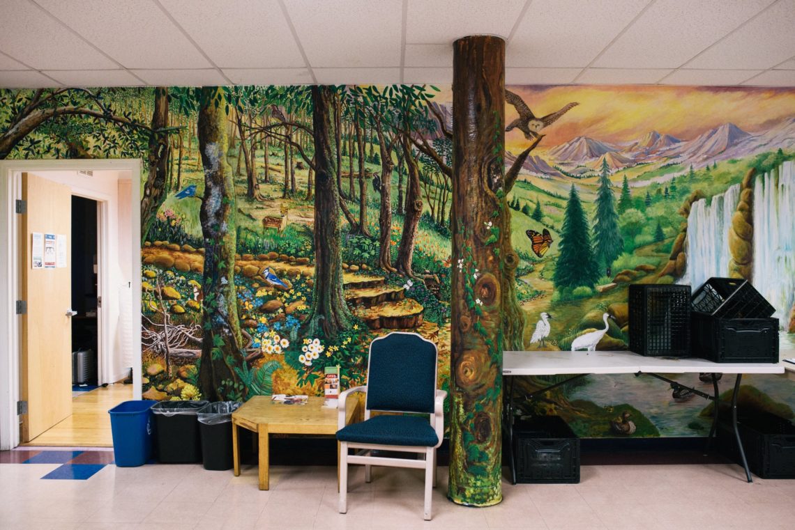 A room with a vibrant wall mural depicting a forest scene, including trees, a waterfall, animals, and mountains. There's a chair, a small round table with a puzzle, and recycling bins. A door to another room is open on the left, hinting at where Berkeley Journalism students might find inspiration.