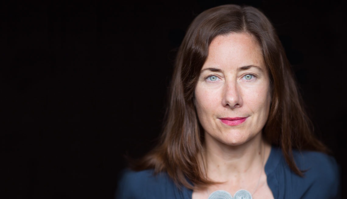 A woman with long brown hair and blue eyes is shown against a black background, wearing a dark blue top. With a neutral expression, she looks directly at the camera and two microphone heads are faintly visible at the bottom center of the image, suggesting an interview for Berkeley Journalism.