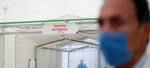 A man wearing a blue face mask stands in the foreground. Behind him, a sign reads "TERAPIA INTENSIVA" above the entrance to a medical facility. Another person is visible in the background, also wearing a mask. The setting appears clinical, likely a COVID-19 intensive care unit captured by Berkeley Journalism.