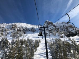 A picturesque view from a ski lift shows a snowy mountain landscape on a clear day. Snow-covered trees dot the mountainside below, and the bright blue sky is partially lined with wispy clouds. Chairlifts are visible, carrying skiers up the mountain, capturing moments worthy of Berkeley Journalism.