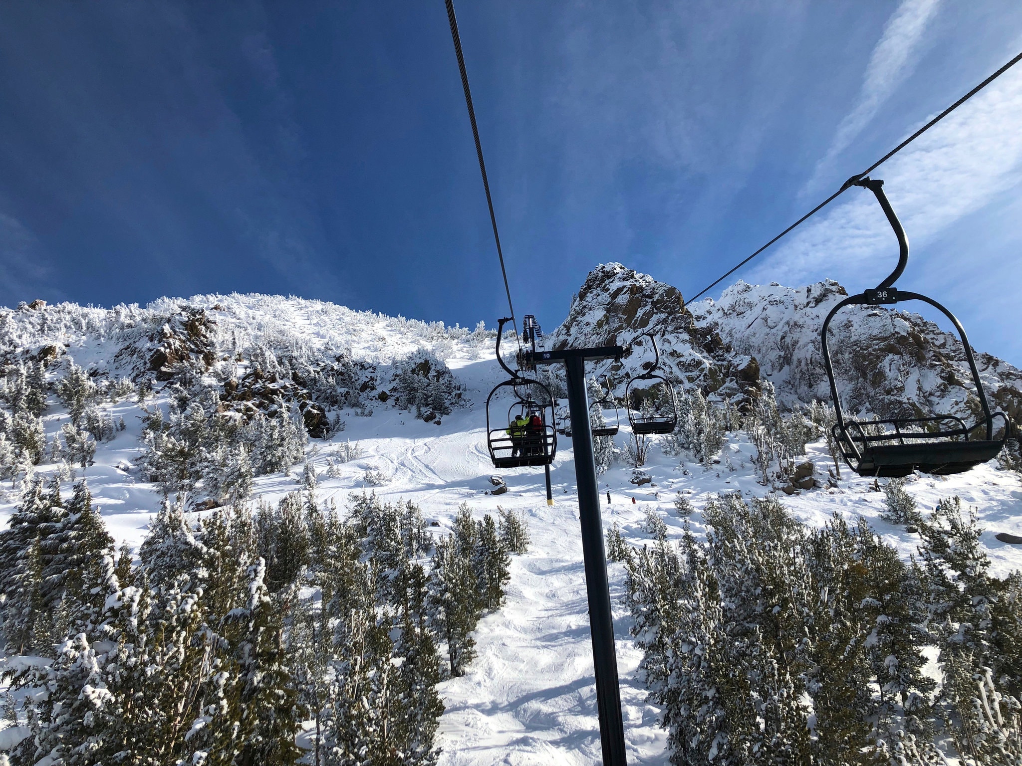A picturesque view from a ski lift shows a snowy mountain landscape on a clear day. Snow-covered trees dot the mountainside below, and the bright blue sky is partially lined with wispy clouds. Chairlifts are visible, carrying skiers up the mountain, capturing moments worthy of Berkeley Journalism.
