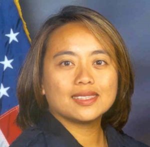 A woman with shoulder-length brown hair and a slight smile is shown in a close-up photograph. She is standing in front of an American flag which is partially visible in the background, embodying the spirit of Berkeley Journalism with her poised presence.