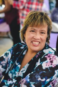 A woman with short hair and a floral-patterned top smiles at the camera. She is seated at what appears to be an outdoor gathering or event, likely one associated with Berkeley Journalism, with other people and blurred colorful backgrounds behind her.