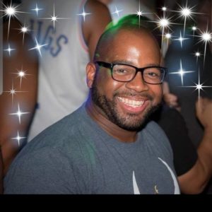 A man with glasses and a beard is smiling broadly while wearing a grey t-shirt. There are sparkles superimposed on the photo, giving it a celebratory or festive feel. A blurred figure in a white jersey is seen in the background, capturing the vibrant spirit of Berkeley Journalism.