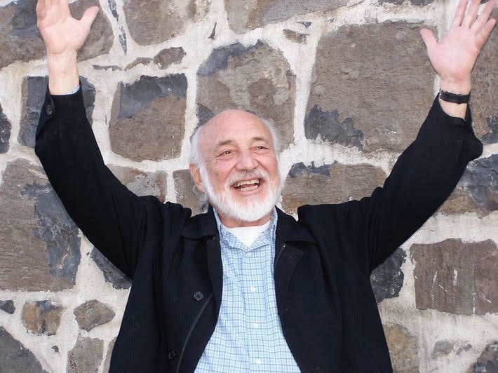 An elderly man with a white beard and mustache is smiling broadly with both arms raised in the air, exuding a youthful enthusiasm. Wearing a black jacket over a light blue shirt, he stands proudly in front of a stone wall, reminiscent of the dynamic storytellers from Berkeley Journalism.