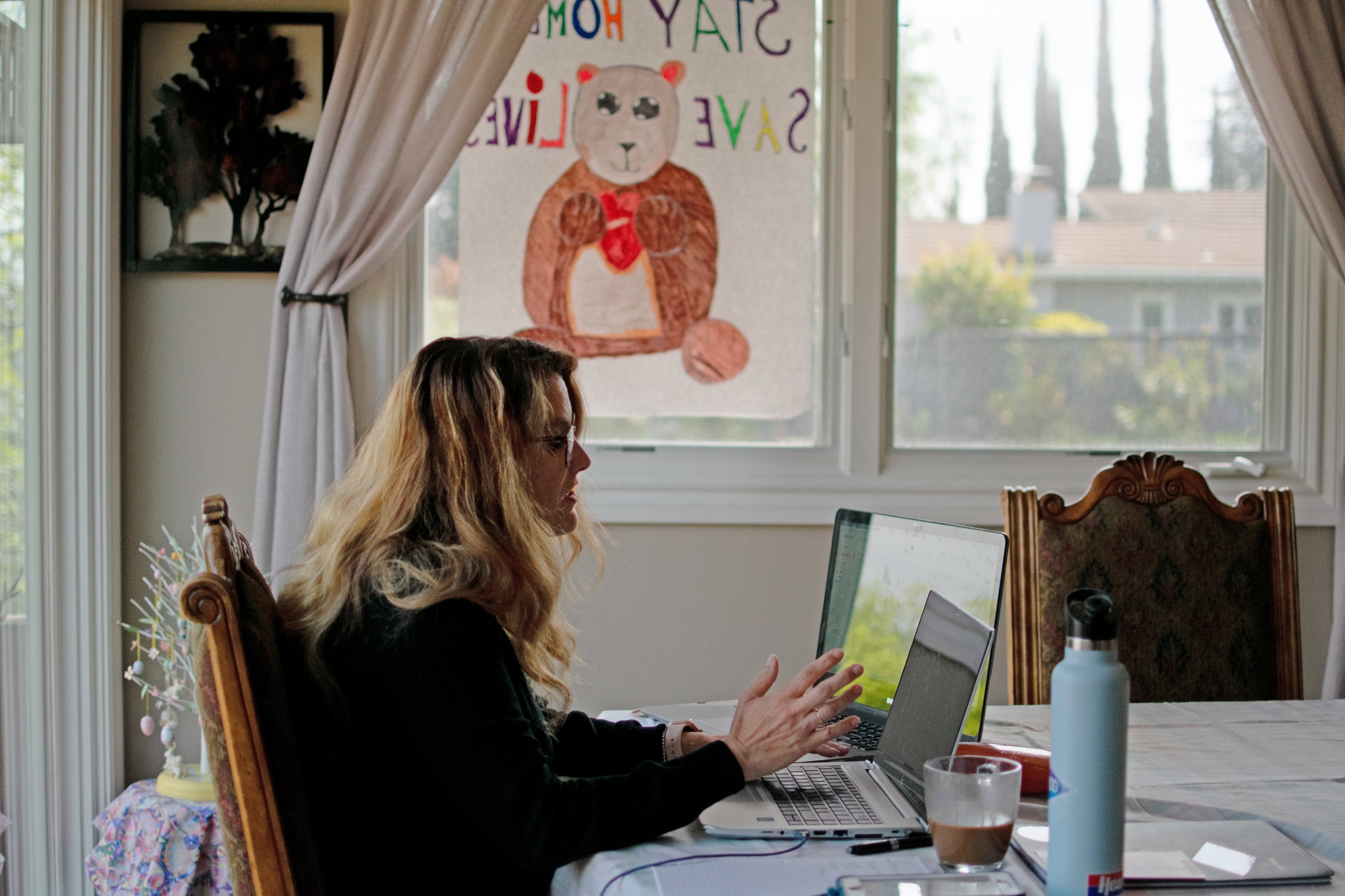 A woman is sitting at her dining room table doing work on her laptop. She has coffee and a water bottle beside her. The background consists of a window where you can see outside and children's drawing of a bear next to to the window. 