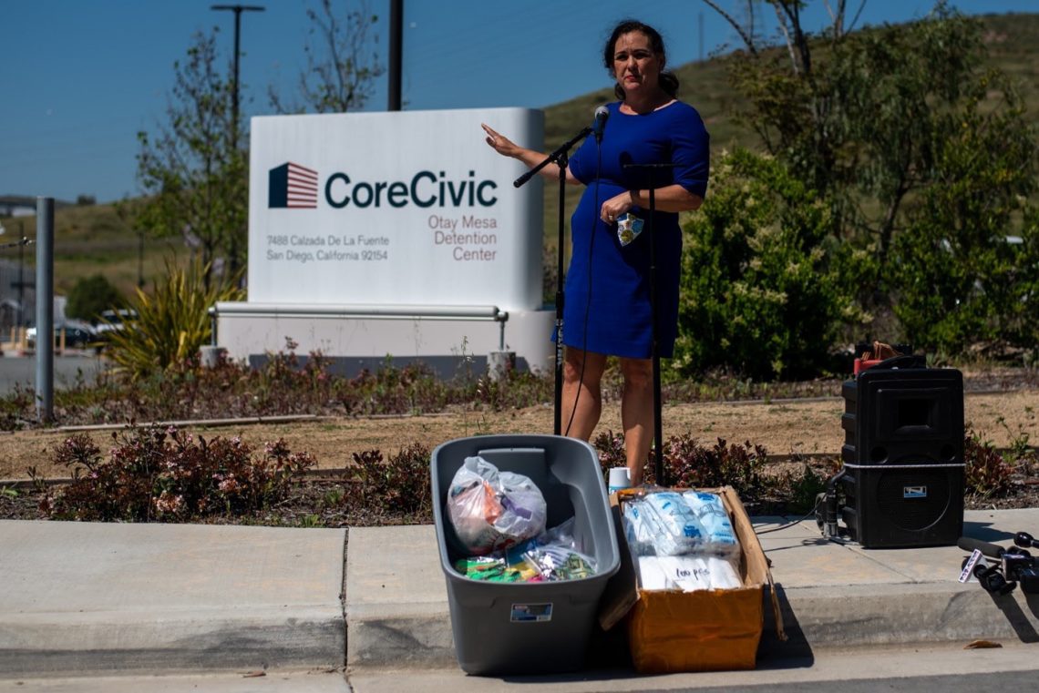 A woman in a blue dress stands at a podium with a microphone, gesturing with her left hand. She is outside the CoreCivic Otay Mesa Detention Center, which is visible in the background. A container of various supplies is placed on the ground next to her, as Berkeley Journalism students capture the moment.