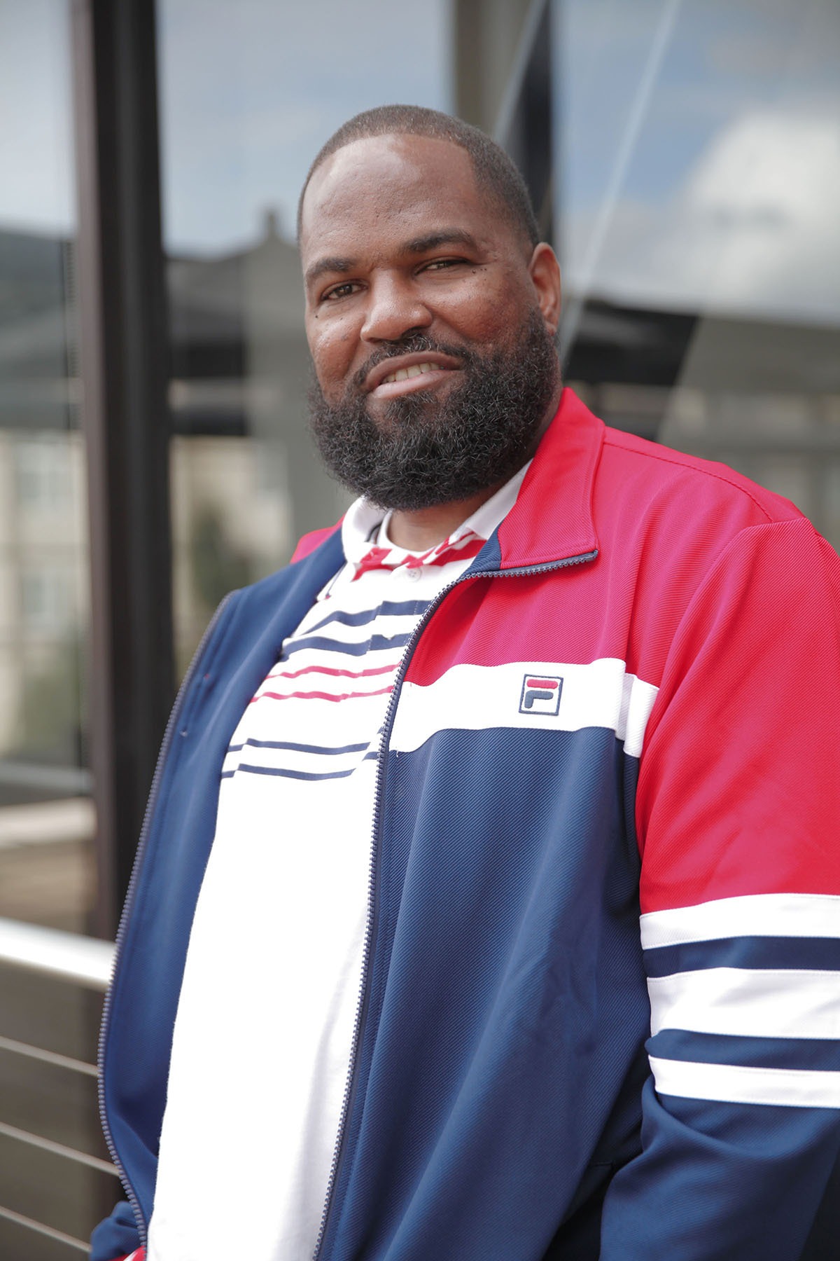 A man with a beard stands outdoors, wearing a red, blue, and white track jacket and a matching striped shirt. He is smiling slightly and looking at the camera. The background shows a modern building with large windows, exuding an atmosphere reminiscent of Berkeley Journalism.