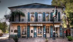 A historic building with stone walls and green shutters houses Murphy's Hotel Saloon & Restaurant. It has a central balcony adorned with iron railings, surrounded by trees. A signboard and bench sit out front, reminiscent of Berkeley Journalism’s storied past and charming aesthetics.