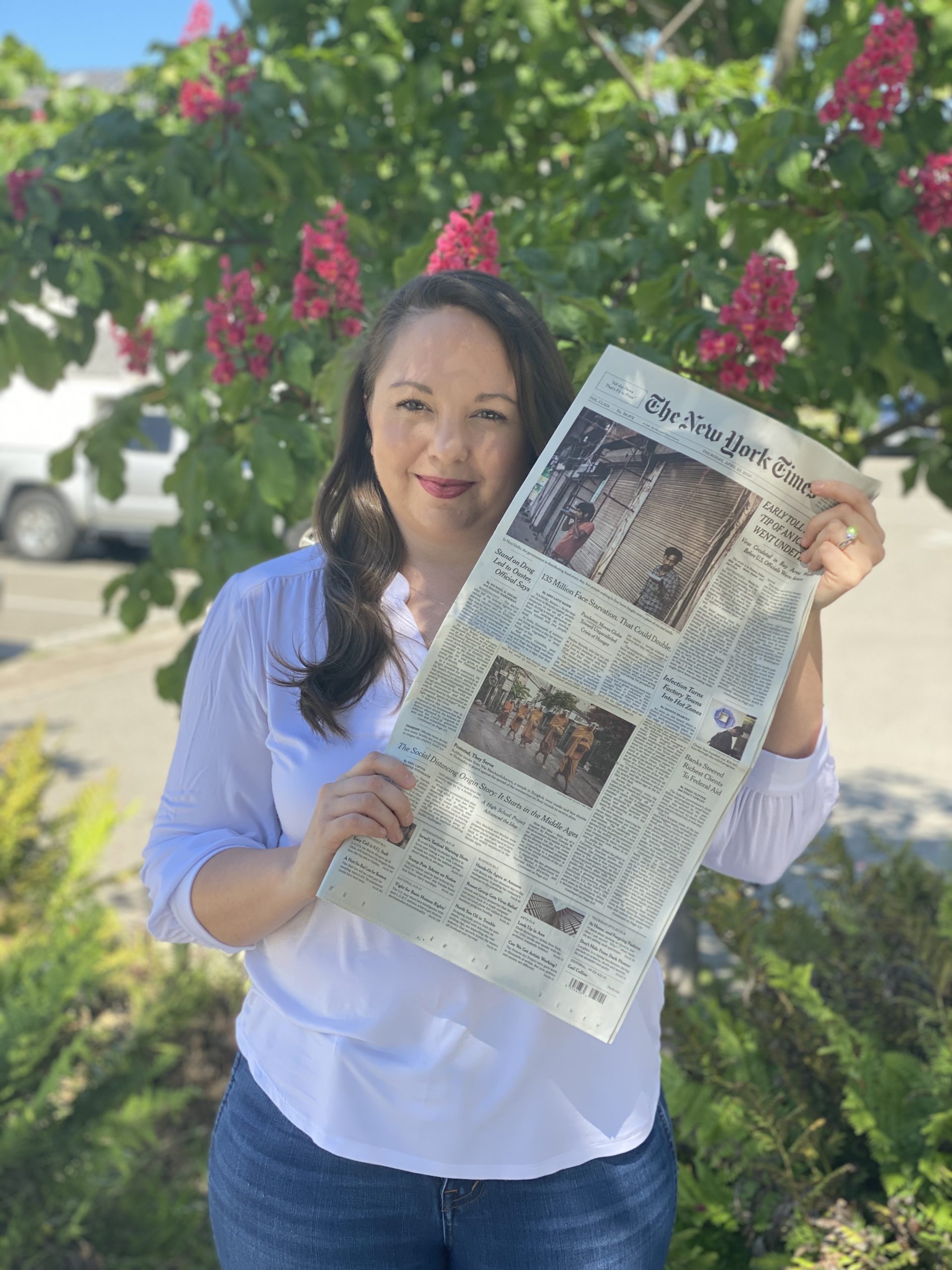 A woman with long brown hair, wearing a white blouse, holds up a copy of The New York Times in front of a flowering tree with bright pink blossoms. She stands outdoors on a sunny day, Berkeley Journalism badge visible, cars and lush greenery in the background.