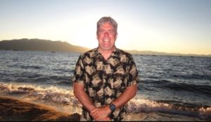 A man stands smiling on a beach during sunset, wearing a patterned shirt. Behind him, gentle waves roll onto the sandy shore, and mountains are visible in the distance. The sky is clear, with soft pastel hues from the setting sun—a scene worthy of a Berkeley Journalism cover.