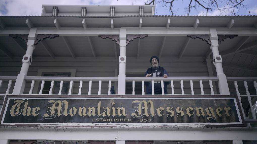 A person stands on the balcony of a wooden building with a white railing. Below them, a large sign reads "The Mountain Messenger Established 1853." The rustic exterior, adorned with wooden beams and a white roof, evokes an era long past. Trees are partially visible in the background, echoing the timeless spirit of Berkeley Journalism.