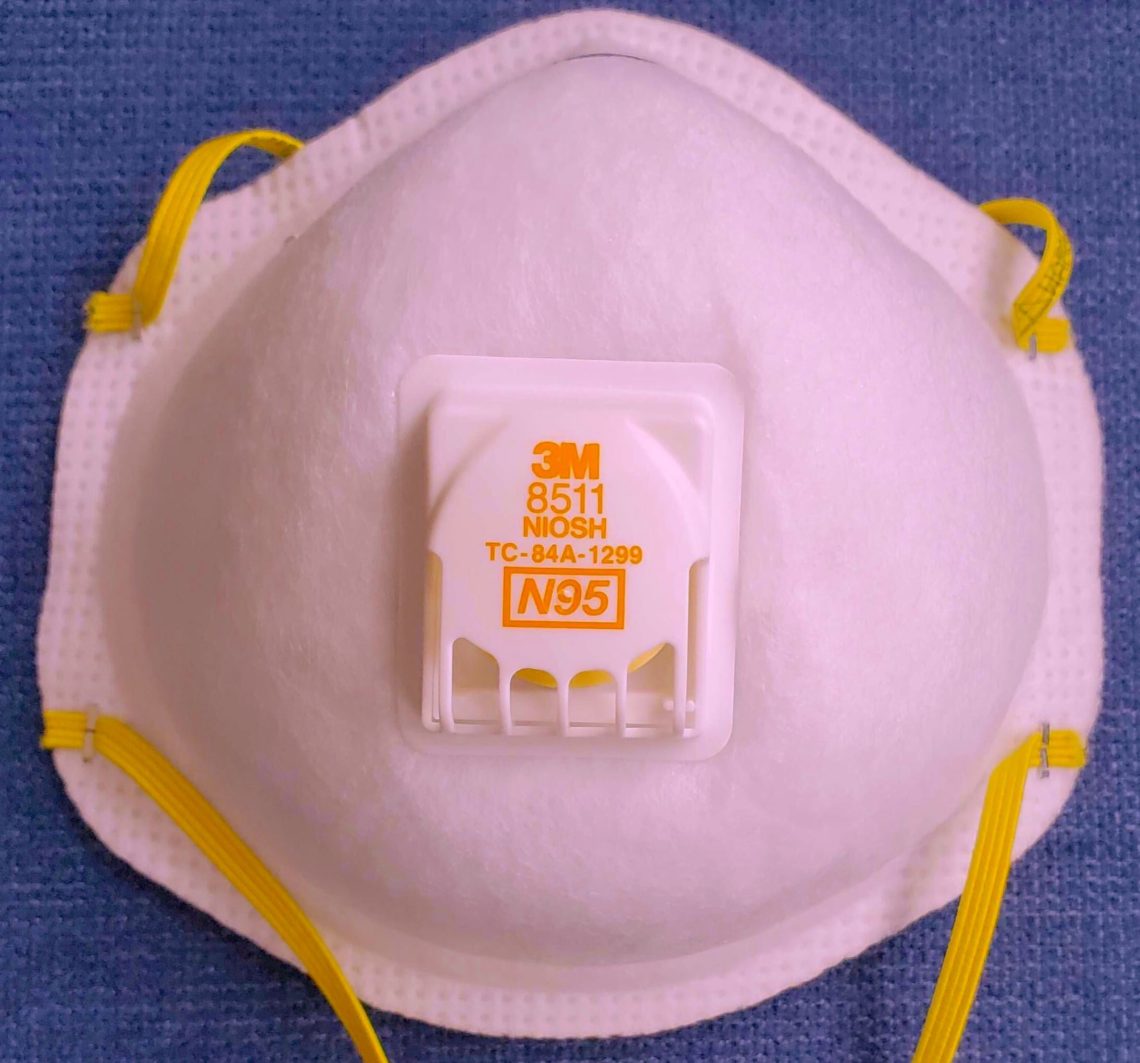 An N95 respirator mask with yellow straps and a white front, perfect for the bustling Berkeley Journalism scene. The mask features a white breathing valve in the center, labeled "8511" with yellow text indicating "3M" and "NIOSH." It is placed on a blue background.