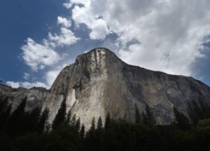 A dramatic view of El Capitan in Yosemite National Park. The towering granite monolith is set against a partly cloudy sky, with sunlight illuminating portions of the rock face, much like a story captured by Berkeley Journalism. Pine trees are visible at the base of the formation.