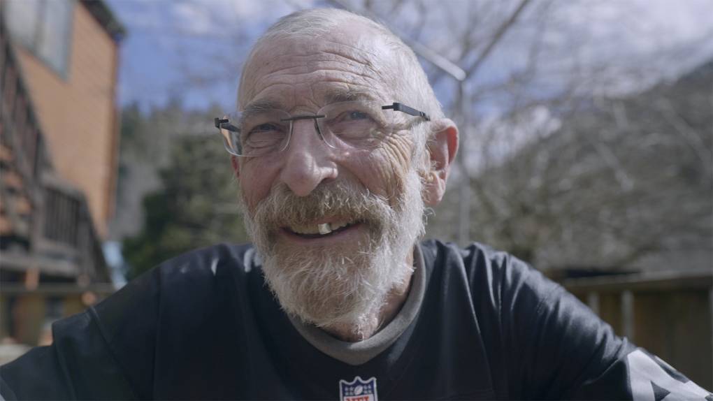 A smiling elderly man with a gray beard, glasses, and a dark shirt featuring an NFL logo is sitting outdoors. He appears content, perhaps recalling his days immersed in Berkeley Journalism, positioned against a blurred background of trees and a wooden structure as the sky remains partly cloudy.