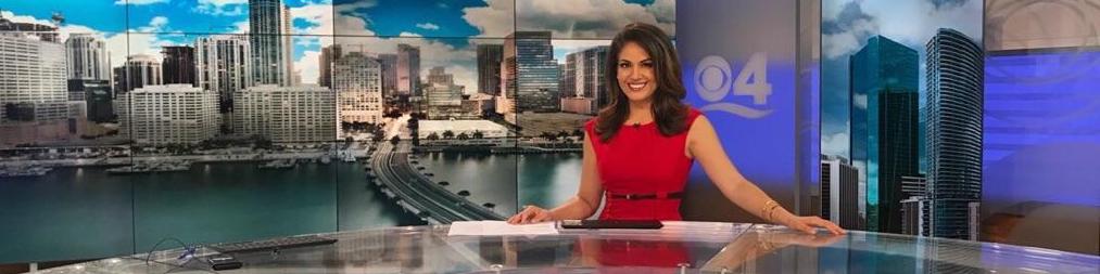 A news anchor in a red dress sits at a glass desk with a cityscape backdrop featuring tall buildings and a bridge over water. The number 4 and a wave symbol are visible on the wall behind her, suggesting it is a Berkeley Journalism studio.
