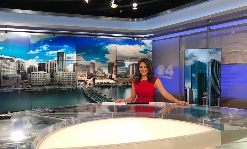 A woman in a red dress sits at a news desk in a modern Berkeley Journalism broadcast studio. Multiple monitors behind her display a city skyline with tall buildings and a bridge over water. Studio lighting illuminates the set, giving it a professional ambiance.