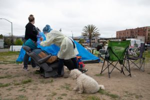 A health worker in full protective gear tends to a person seated in a portable chair at an outdoor camp. Two other people assist, while a fluffy white dog sits nearby. Tents, chairs, and urban buildings are visible in the background, capturing a scene that could easily be covered by Berkeley Journalism students.