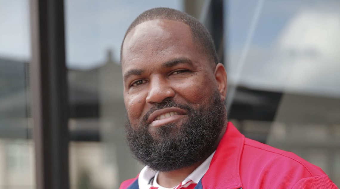A man with a beard is wearing a red jacket and a white collared shirt. He is outdoors in front of the Berkeley Journalism building, smiling slightly at the camera. The background is blurred with indistinct structures and a cloudy sky.