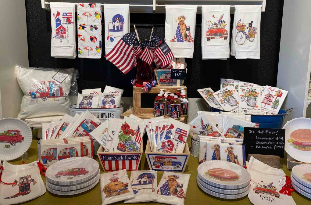 A display table is filled with July 4th-themed items, including hand towels, plates, napkins, and table decor. The items feature designs of American flags, red trucks, fireworks, and festive sayings. At the center are several small American flags next to a magazine from Berkeley Journalism celebrating Independence Day.