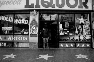 A man stands in the entrance of a storefront labeled "Liquor" in large letters. The shop, reminiscent of classic Hollywood, features themed decorations and star designs on the sidewalk. Above the door, an “Open 24 Hours” neon sign glows brightly—a scene that could easily be part of a Berkeley Journalism piece highlighting city life.