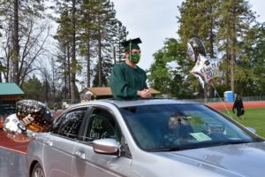 A graduate in a green cap and gown stands through the sunroof of a car decorated with balloons that read "Class of 2020". The car, bearing signs for Berkeley Journalism, is on a track with trees and a field in the background. Another person sits inside the car.