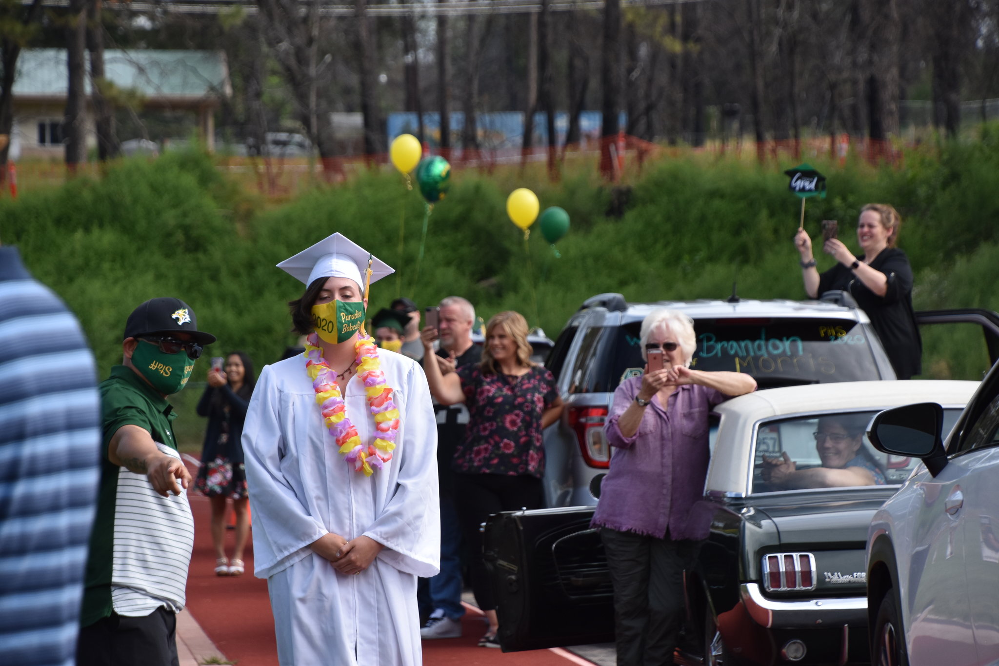 A graduate in a white cap and gown adorned with a pink lei stands on a track. People, some wearing masks, celebrate nearby. Cars are parked along the track, with onlookers cheering, holding balloons and signs. One sign reads 
