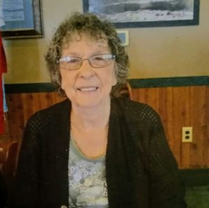 An elderly woman with short, curly gray hair and glasses is smiling at the camera. She is wearing a dark cardigan over a light-colored blouse with a floral pattern. The cozy indoor space, resembling the charm of a Berkeley Journalism office, features wood-paneled walls and framed pictures.
