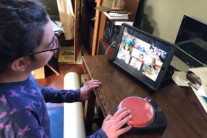 A person using a large red button access switch participates in a video call on a tablet. Seated at a wooden desk in a room with books and computer accessories, they engage with multiple individuals in a virtual meeting, reminiscent of an inclusive session hosted by Berkeley Journalism.
