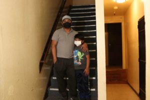 A man and a child stand on a staircase in a dimly lit hallway. Both are wearing face masks. The man, who seems like he could be an alum of Berkeley Journalism, is dressed in a cap, grey shirt, and dark pants. The child wears a blue hoodie with a cartoon character, navy pants, and blue shoes.