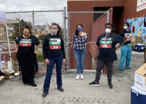 A group of five people stands outdoors near a chain-link fence. Three individuals are wearing "United In Health D-10" shirts, all donning face masks. Boxes and a cooler are nearby. The background includes a mural on a building wall, capturing the essence of Berkeley Journalism's commitment to community stories.