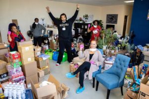 A group of six people are celebrating amidst a room filled with packed boxes and assorted items. Some are raising their arms in triumph, while others are smiling and seated. The background includes potted plants, various storage items, and a poster for Berkeley Journalism.